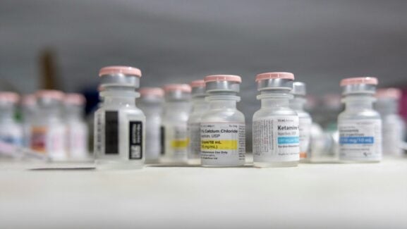 A 10 mL multi-dose ketamine hydrochloride vial on a clinic counter beside other injectable drug vials